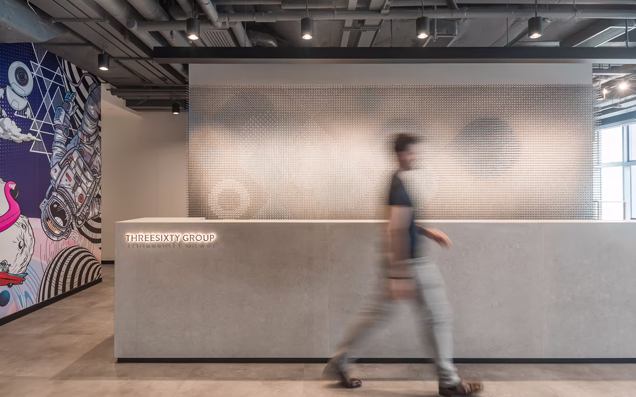Modern office reception area with a concrete desk labeled 'THREESIXTY GROUP', a wall with a textured metallic panel, and a blurred person walking by.