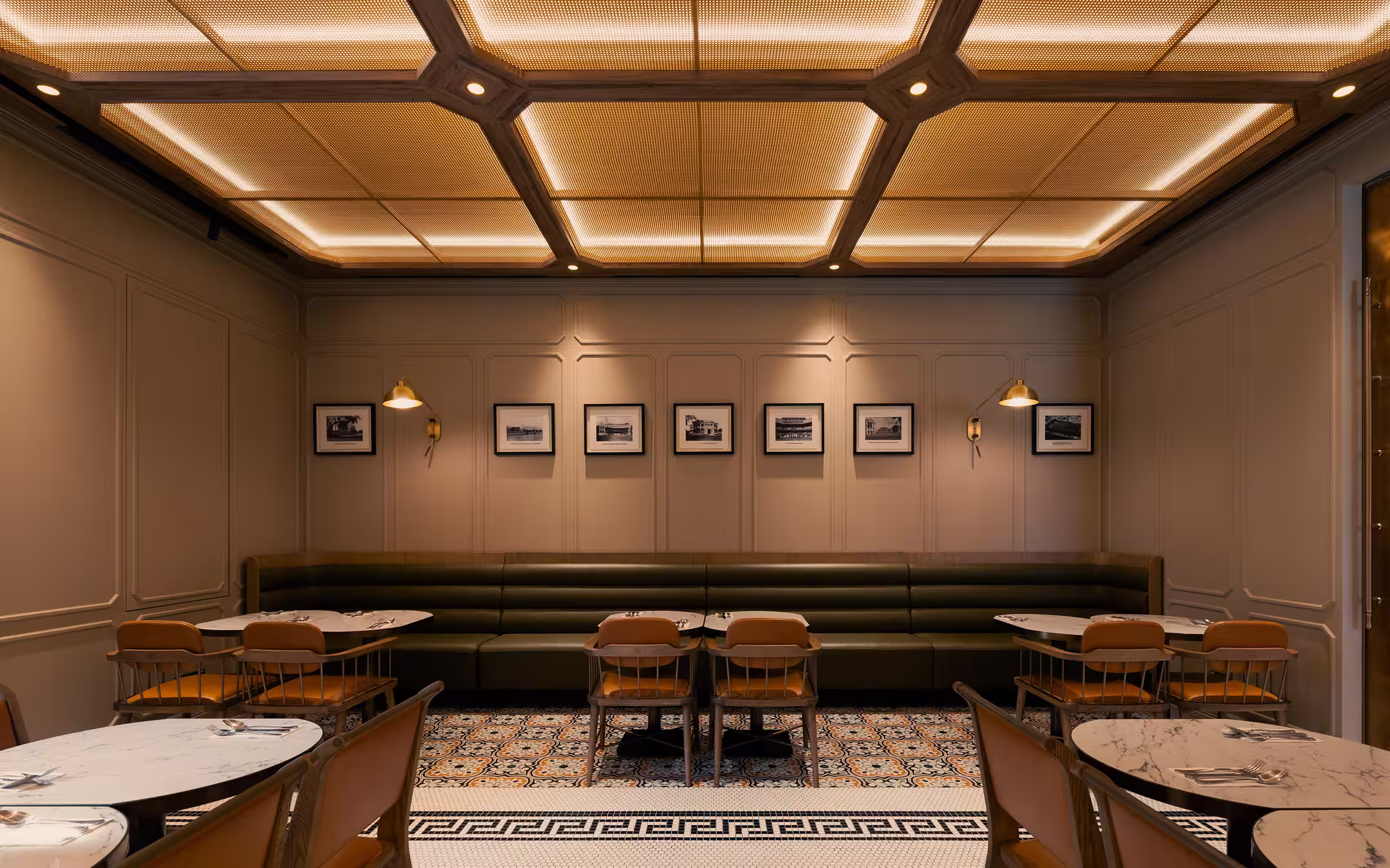 Cozy restaurant interior with marble tables, brown chairs, a long dark green cushioned bench, framed photos on beige paneled walls, and warm overhead lighting.