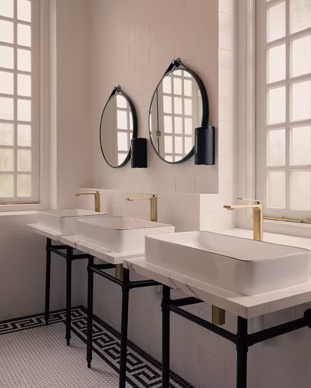 Modern bathroom with three white rectangular sinks on black metal stands, gold faucets, oval mirrors, and large frosted windows.