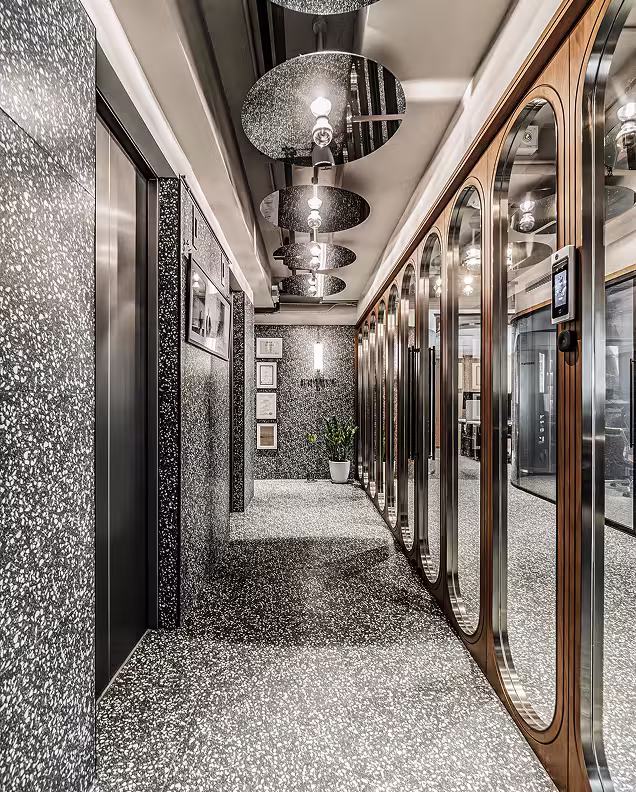 Modern hallway with terrazzo flooring and walls, black elevator door, wood-framed glass partitions, hanging round light fixtures, and a potted plant at the end.