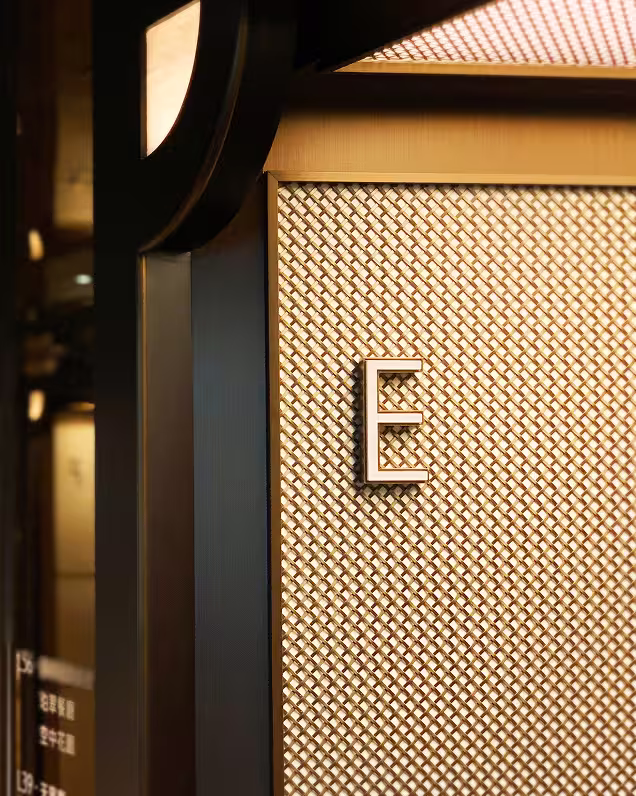 Close-up of a textured wall panel with a raised letter E mounted on it, illuminated by warm lighting.
