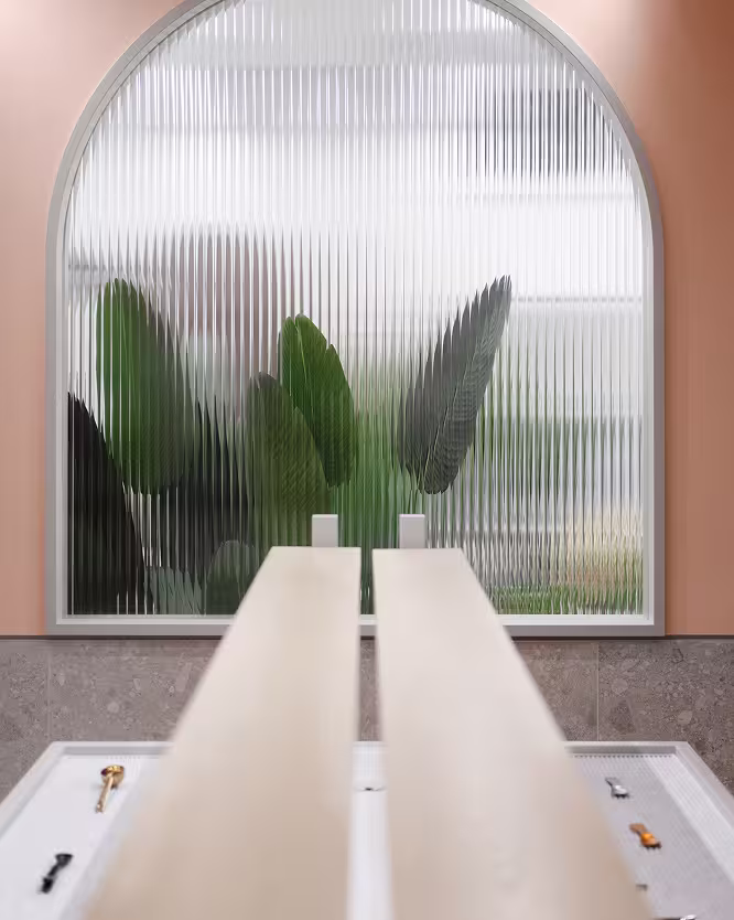 Indoor architectural window with ribbed glass framed by a rounded arch and pink walls with large green leaves blurred behind the glass.