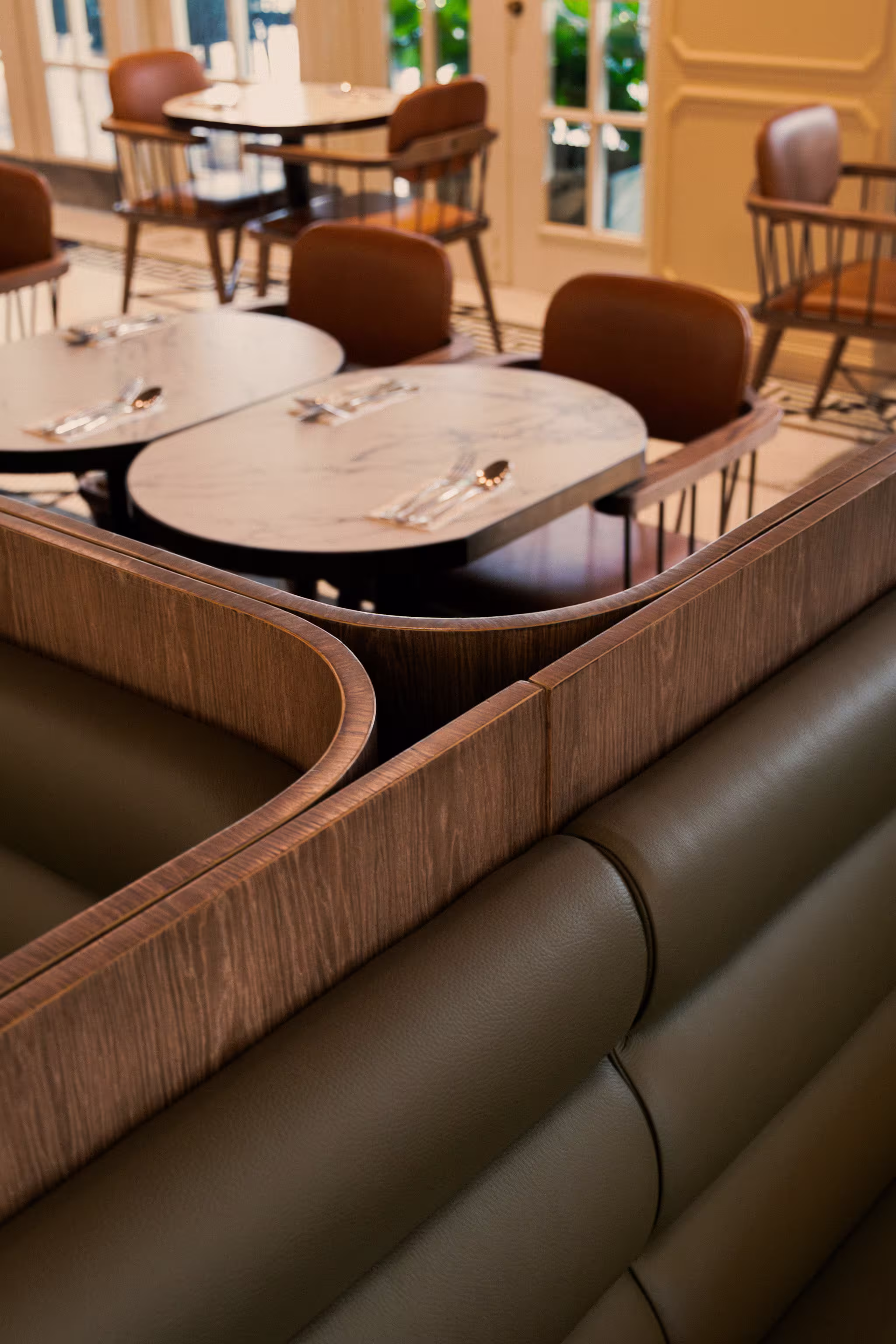 Modern restaurant seating with tufted olive-green leather booth and marble tabletops set with silverware and brown wooden chairs.