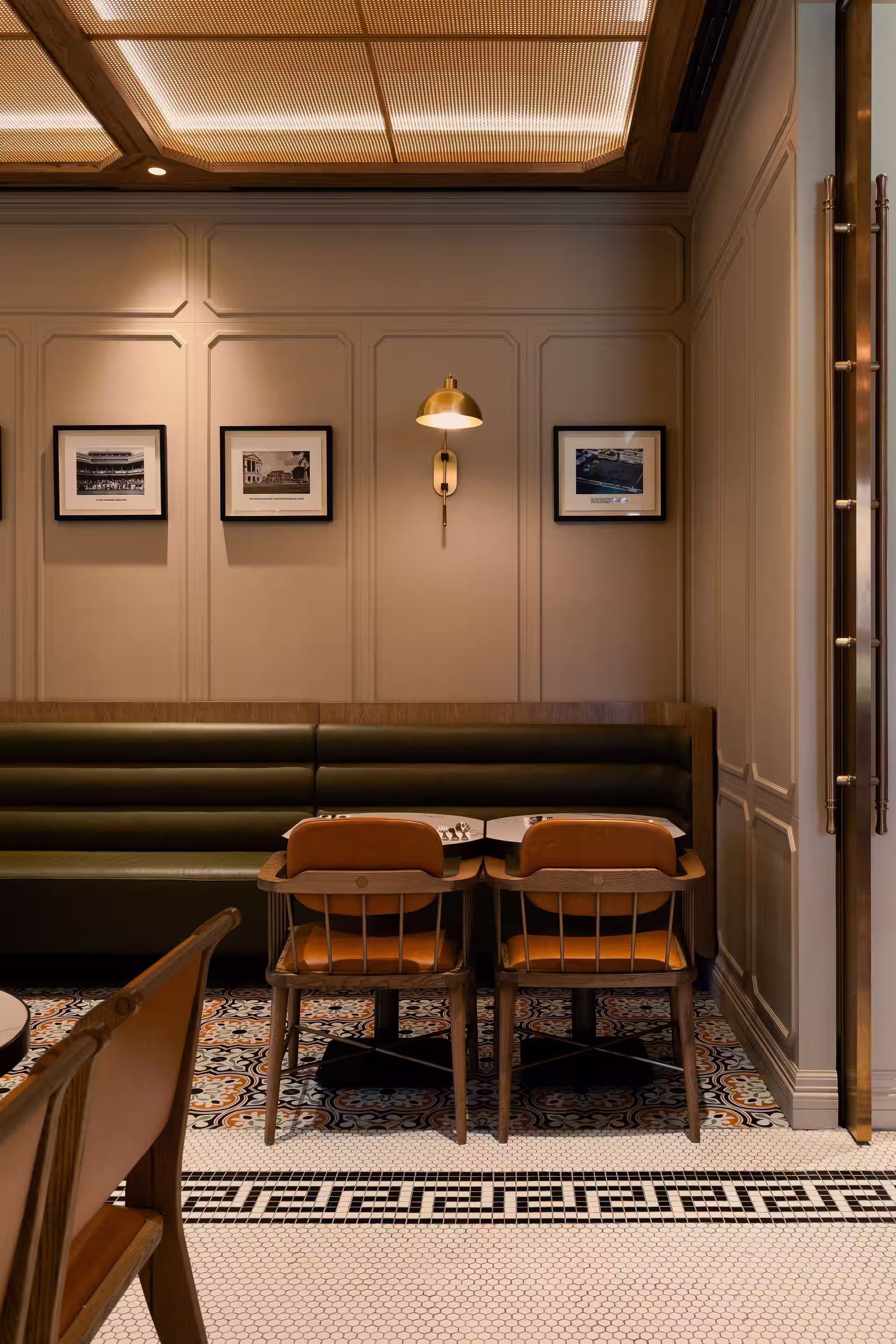 Cozy restaurant corner with two wooden chairs and a green cushioned bench, decorated with framed black-and-white photos on beige paneled walls and patterned tile flooring.