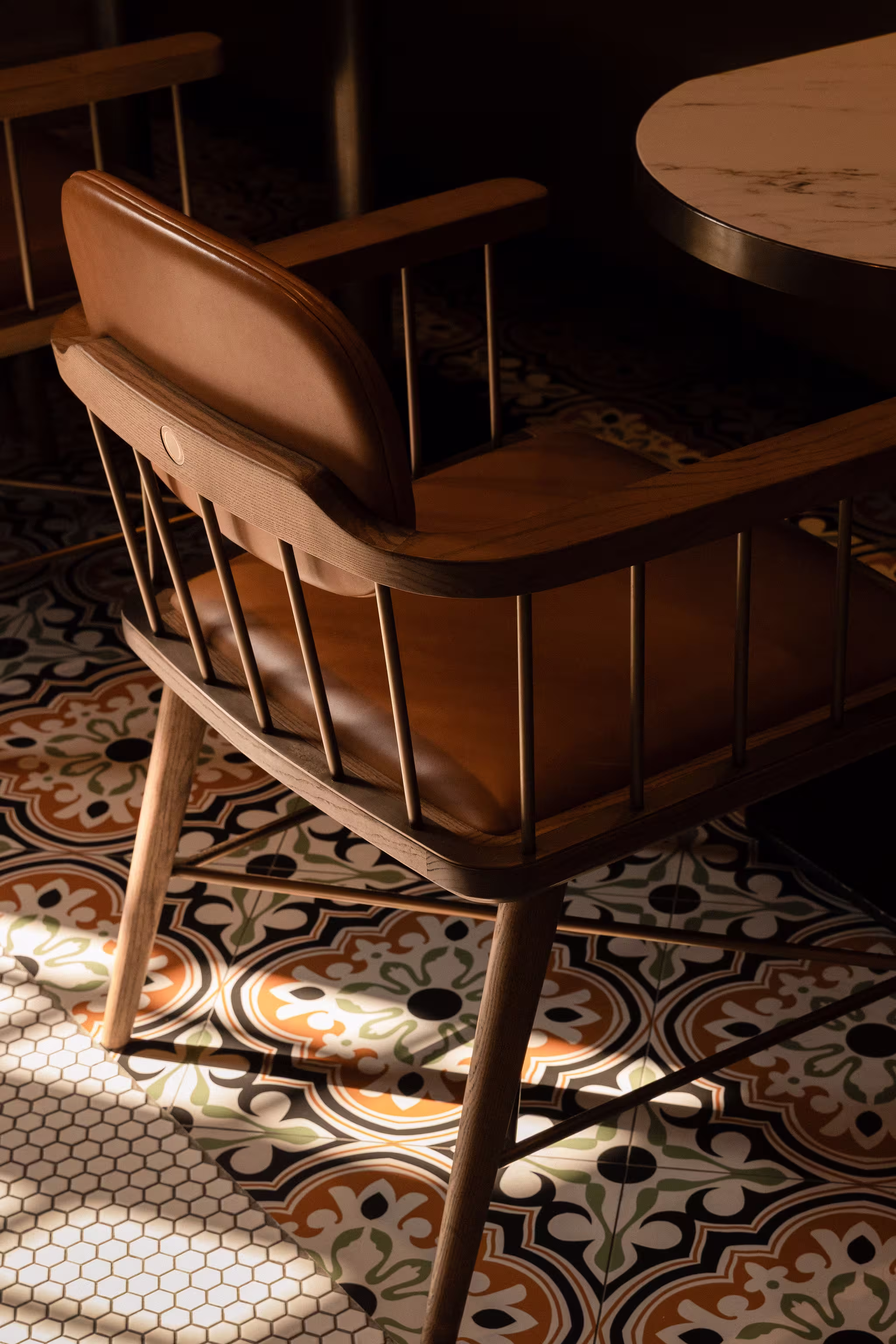 Wooden chair with brown leather cushion and backrest next to round table on patterned tile floor with sunlight and shadows.