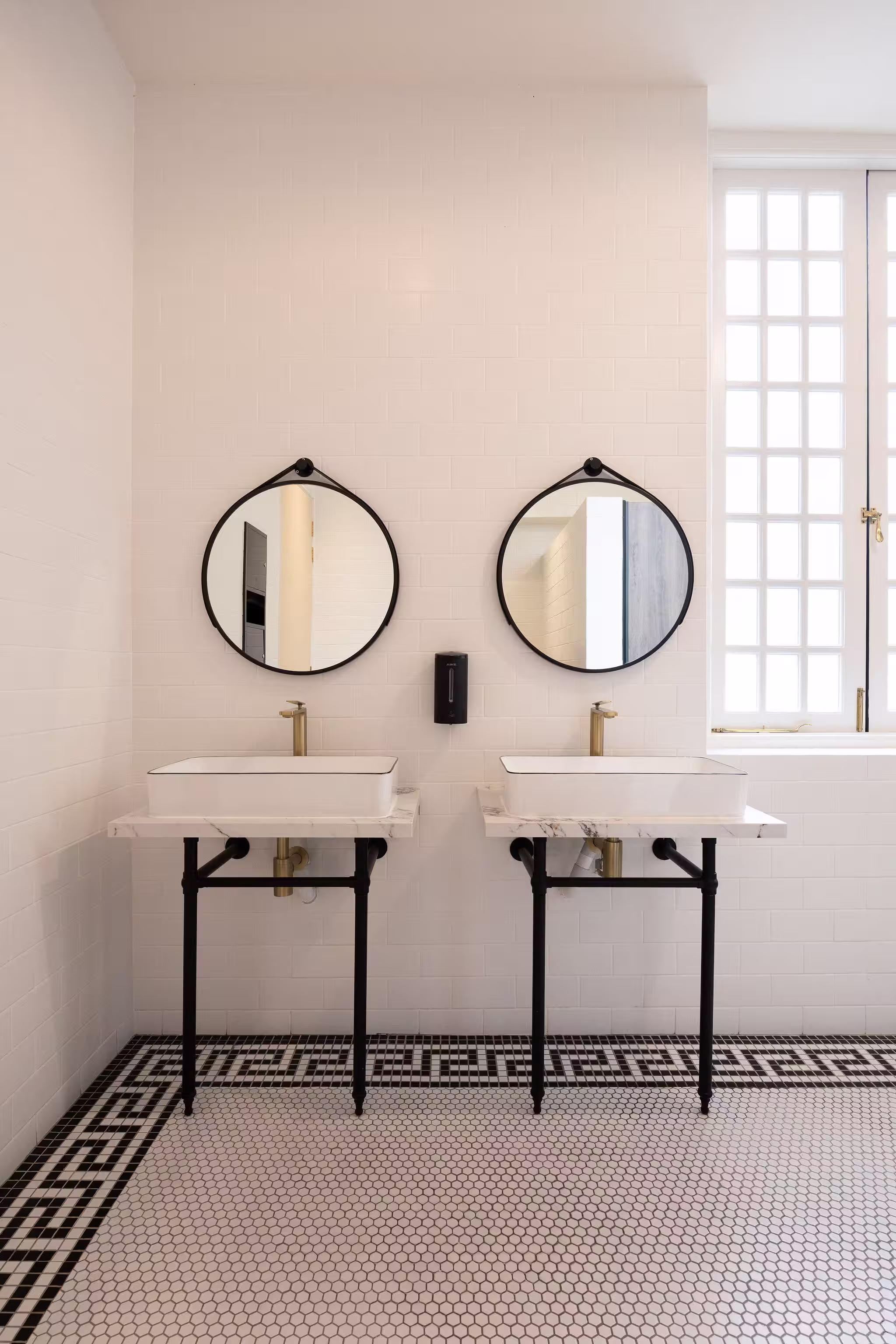 Modern bathroom with two rectangular white sinks on black-legged marble countertops, round black-framed mirrors, white tiled walls, and black and white hexagonal tile floor.