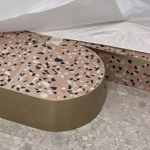 Oval terrazzo table with black, white, and pink speckles on a gold base, next to a terrazzo bench and light-colored speckled floor.