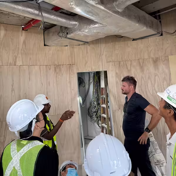 Group of construction workers and a man in black shirt discussing inside a building under construction with exposed ductwork and plywood walls.