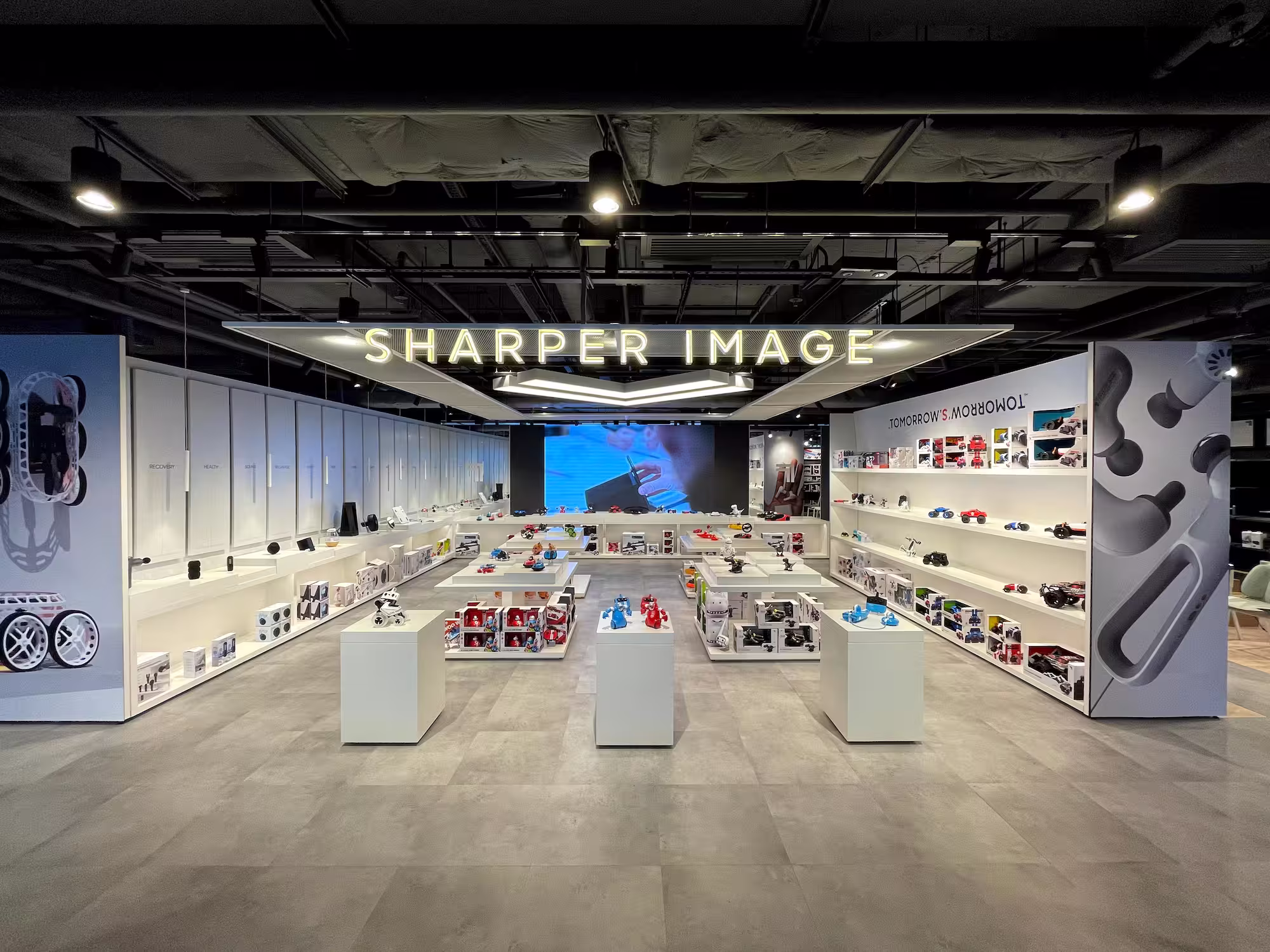 Modern retail store interior with illuminated 'Sharper Image' sign and shelves displaying tech gadgets and robots.