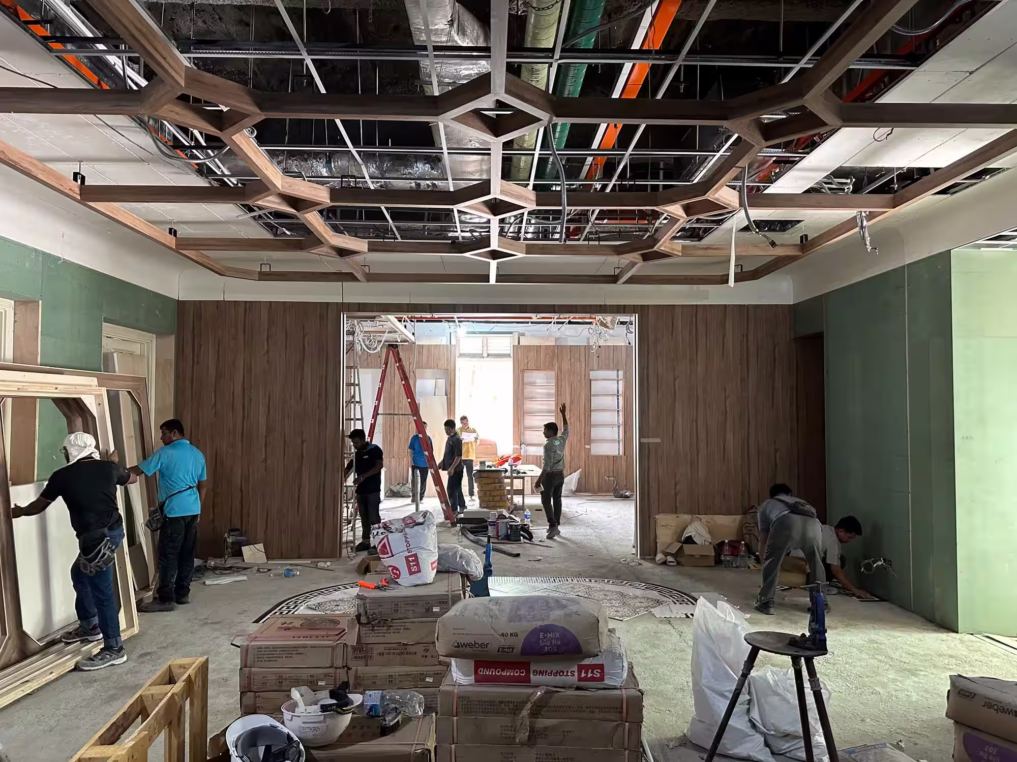 Construction workers installing wood panels and ceiling framework inside a large unfinished room.