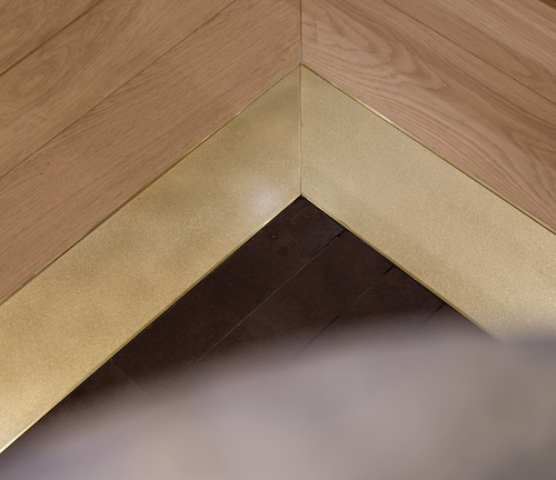 Floor detail where oak herringbone meets darker timber, divided by a chevron of brass inlay
