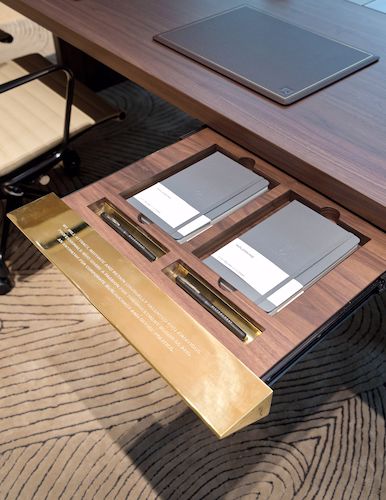 Walnut desk with a brass-lined drawer drawn open, holding notebooks and pens beneath the worktop