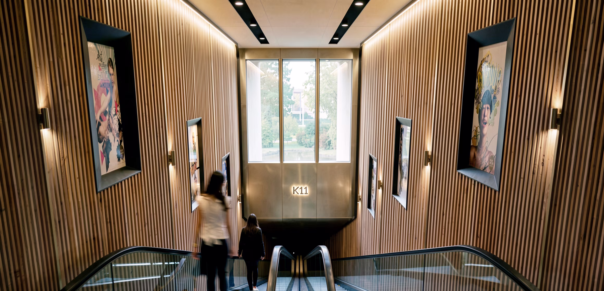 Escalator descending between fluted timber walls hung with artwork, towards the K11 bronze panel