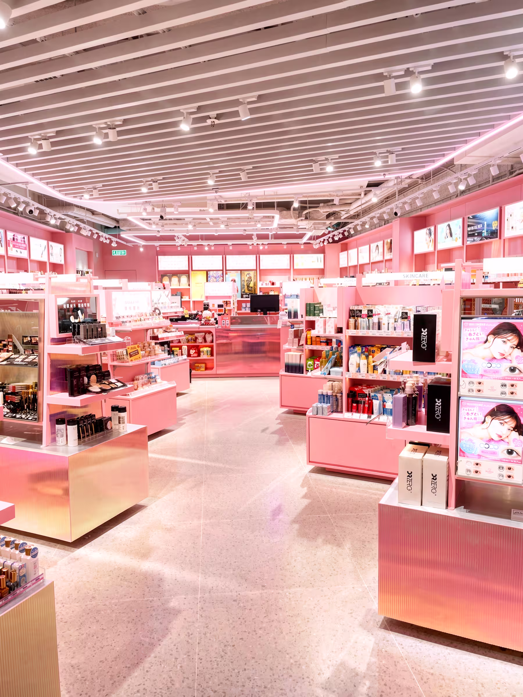 Interior of a pink-toned cosmetics retail concession with display counters and shelving under a slatted ceiling