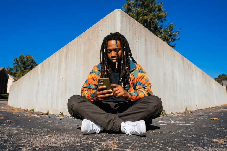 Man sitting cross-legged on asphalt in front of a concrete structure, using a smartphone.