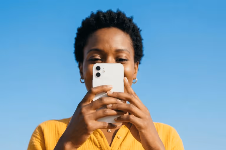 Person holding a white smartphone in front of their face against a clear blue sky