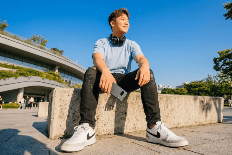 Young man sitting on a concrete ledge outdoors with headphones around his neck, holding a smartphone