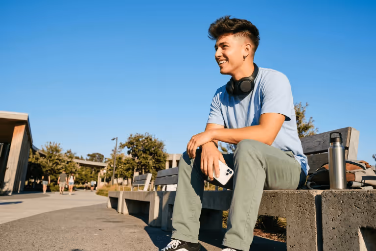 Young man sitting on a bench outdoors, holding a smartphone with headphones around his neck