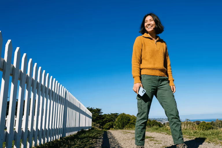 Smiling woman holding a smartphone, standing on a gravel path beside a white picket fence