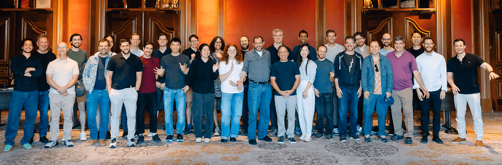 Group of 27 people standing indoors against a wood-paneled wall with red decor, smiling and posing for a group photo.