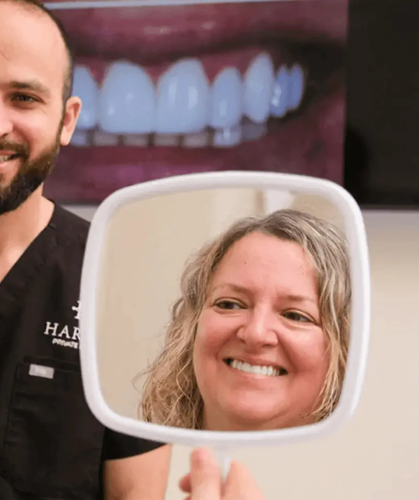Smiling woman looking at her teeth in a handheld mirror with a male dentist standing nearby.