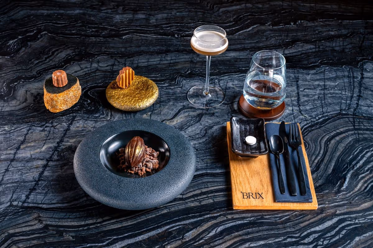 Artfully plated gourmet chocolate dessert with two small pastries, a cocktail glass, a water glass, and black cutlery on a wooden board labeled BRIX, all on a dark textured stone surface.