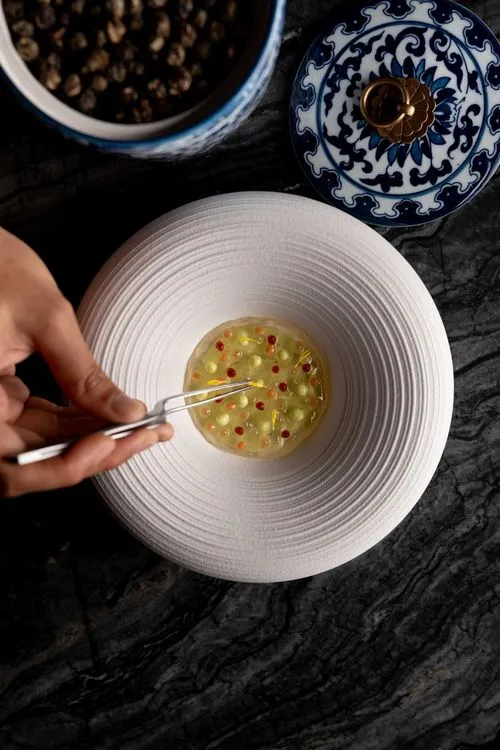 Hand using tweezers to place small colorful spheres in a clear gel on a textured white plate next to a blue and white patterned jar lid.