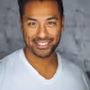 Smiling man with short dark hair wearing a white V-neck shirt against a gray brick wall background.