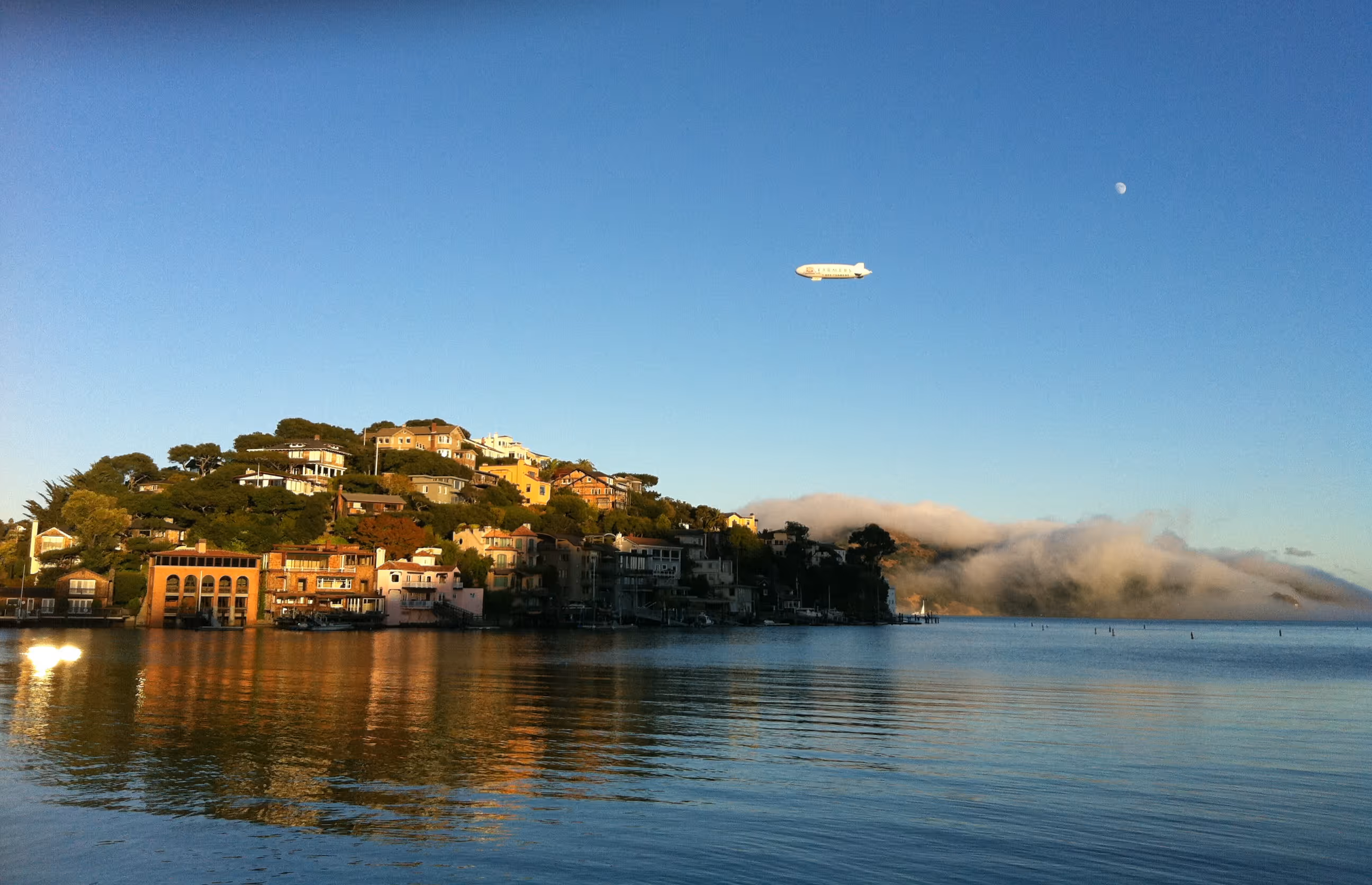 Waterfront houses on a hillside with a blimp flying above and fog rolling over distant hills under a clear blue sky.