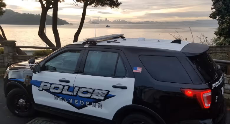 Black and white Belvedere police SUV parked near a waterfront with a city skyline and boat visible in the background at sunset.