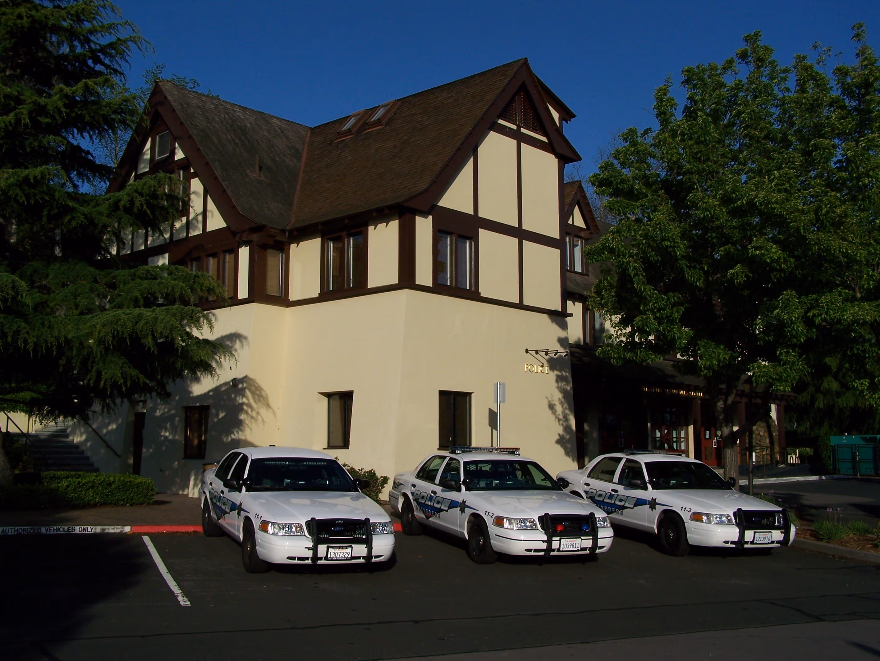 Three white police cars parked in front of a beige and brown Tudor-style building under a clear blue sky.