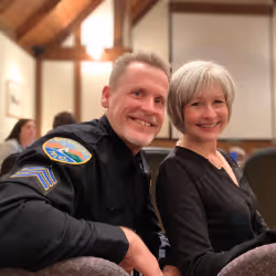 Smiling man in a police uniform sitting next to a smiling woman with short gray hair in an indoor setting.