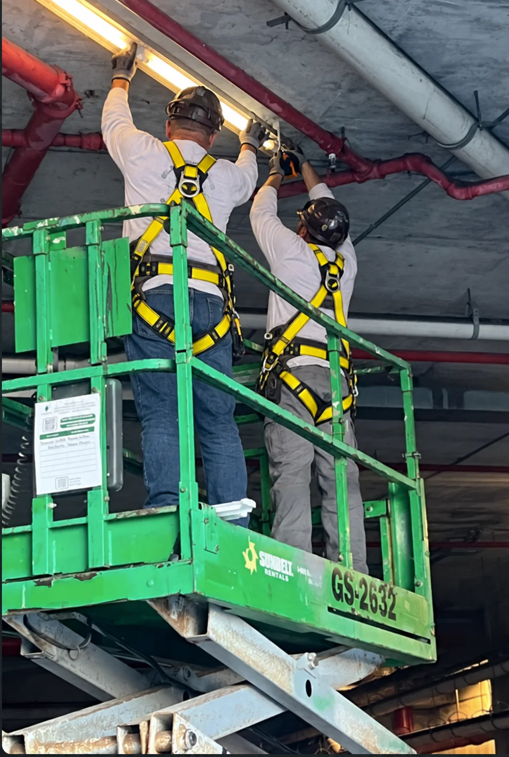 AI-generated illustration of two electricians wearing yellow safety harnesses installing ceiling lighting from a scissor lift
