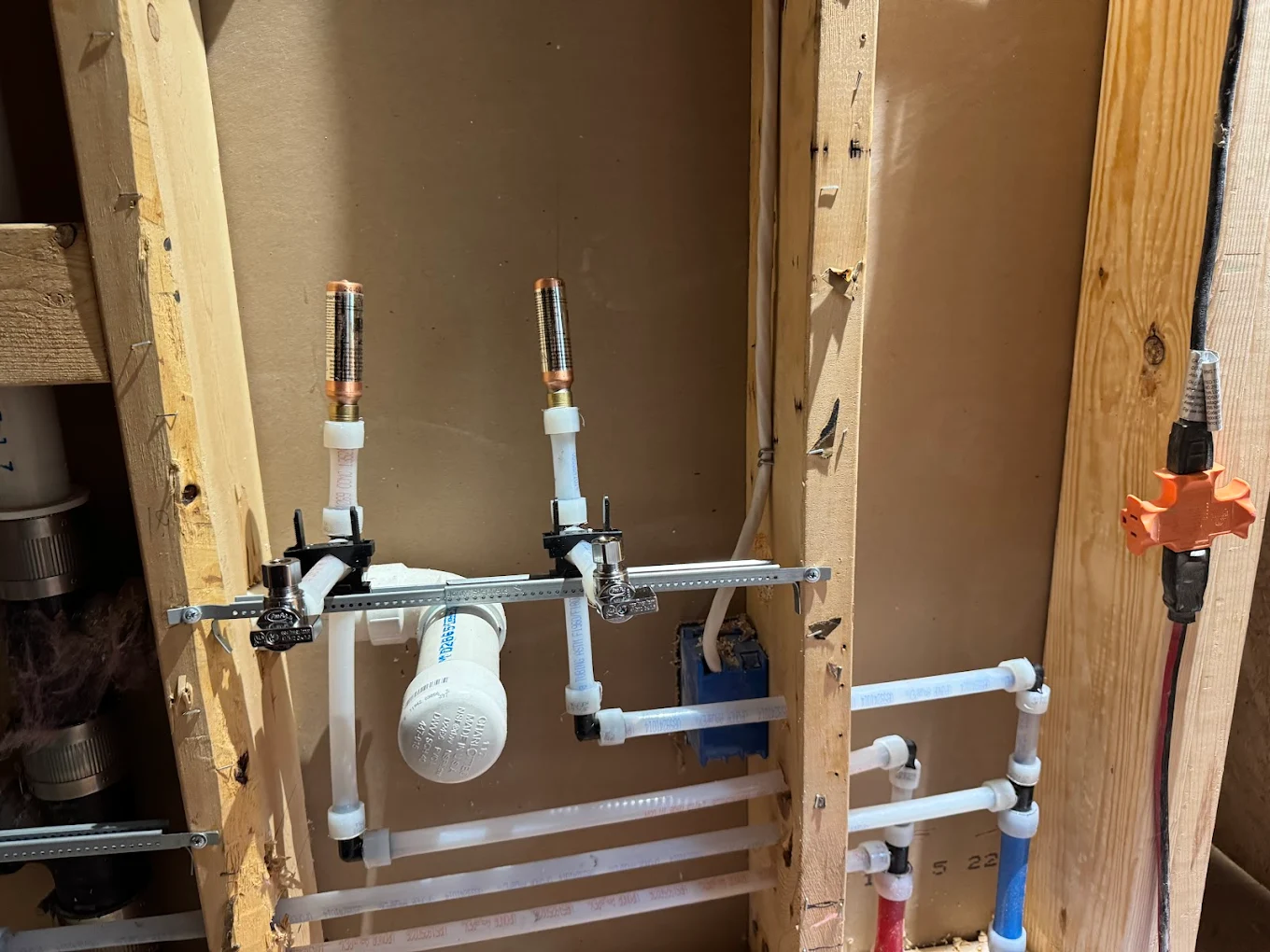 Exposed plumbing pipes and valves inside wooden wall studs with an orange multi-outlet extension cord on the right.
