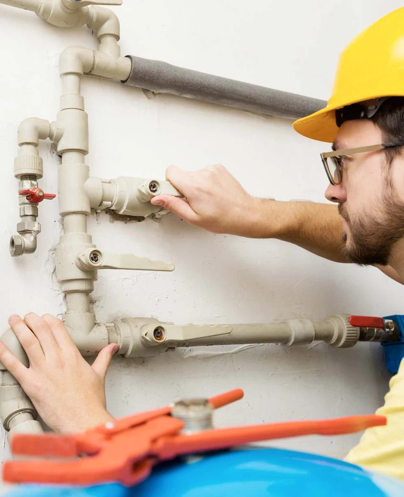 Plumber wearing a yellow hard hat and glasses inspecting white plastic water pipes on a wall.
