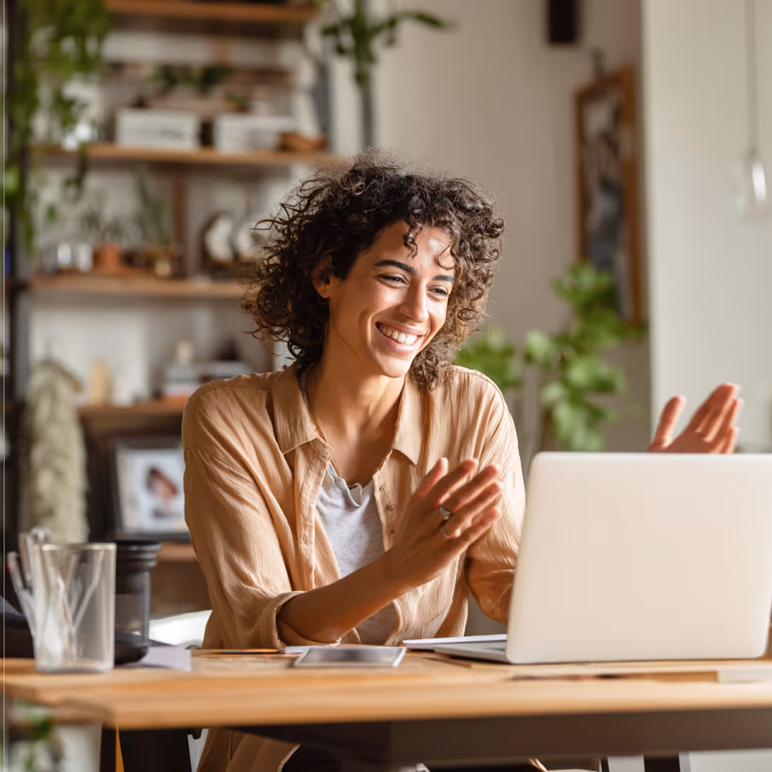 Smiling woman with curly hair video chatting on a laptop at a wooden desk in a cozy room.