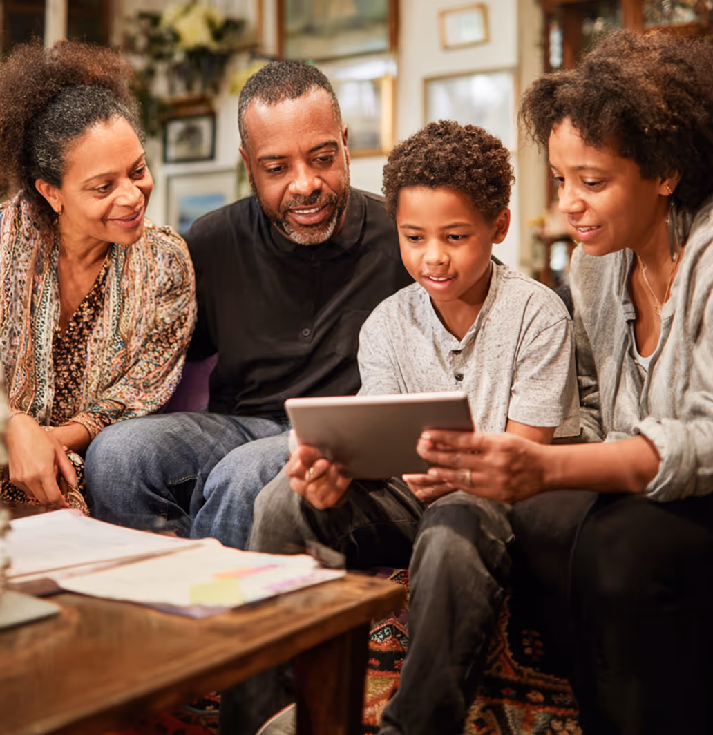 A family of four sitting together on a couch looking at a tablet screen with interest and smiles.