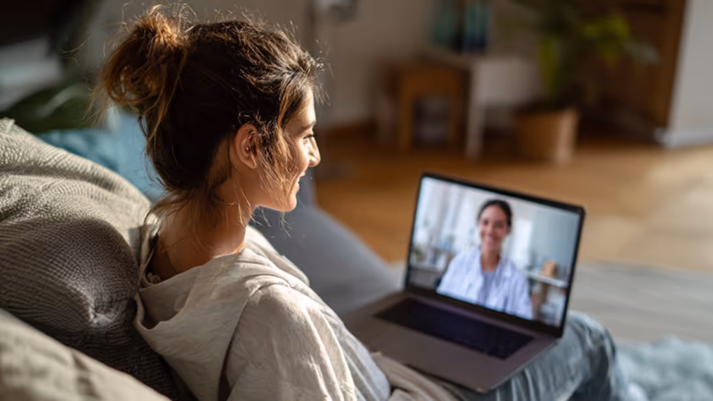 Woman sitting on a couch engaged in a video call with a doctor on a laptop screen.