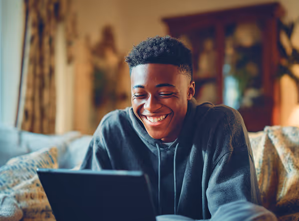 Smiling young man sitting indoors, looking at a laptop screen.