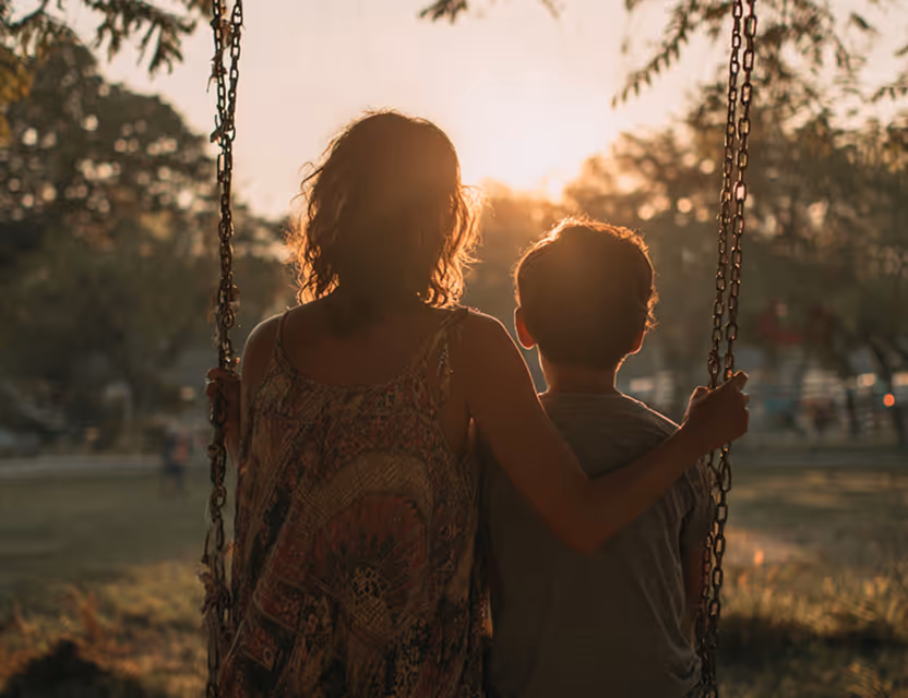 Woman and child sitting together on swings, embracing with sunlight filtering through trees in the background.