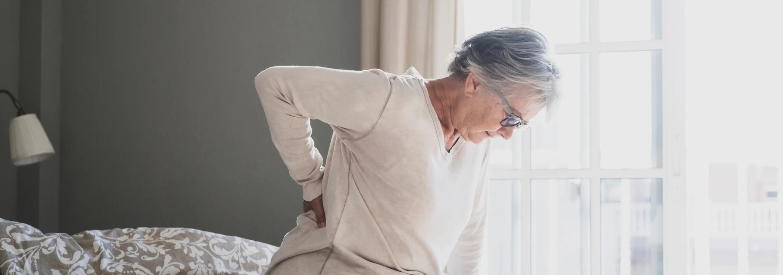 An older woman sitting on a bed with her hand on her lower back