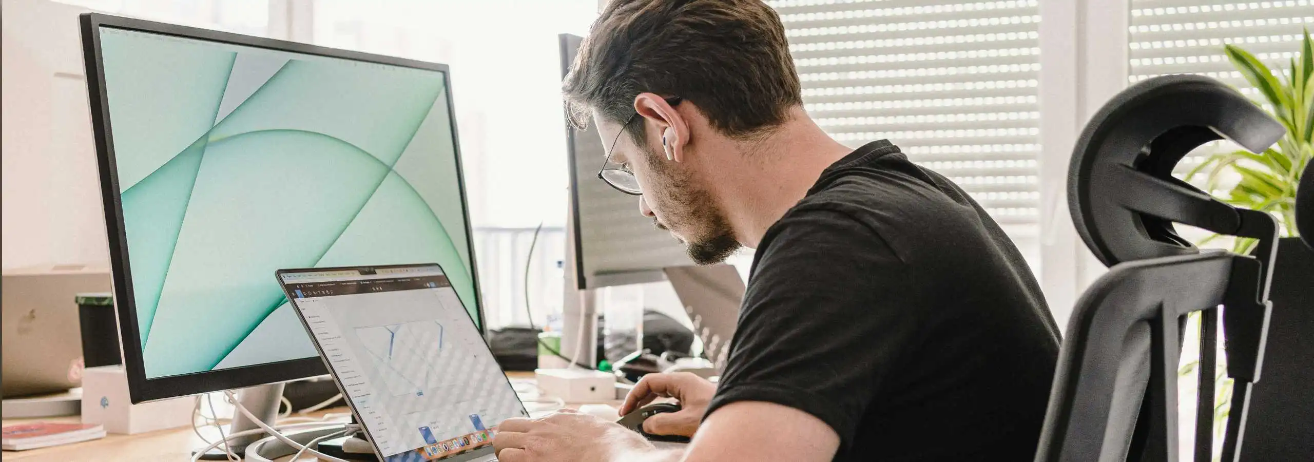 A man leaning forward at a desk, working on a laptop with a large monitor in front of him in a bright office setting.