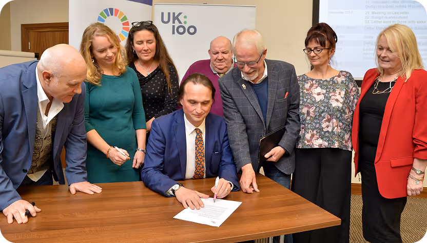 Person in blue suit signs document surrounded by colleagues at UK100 event