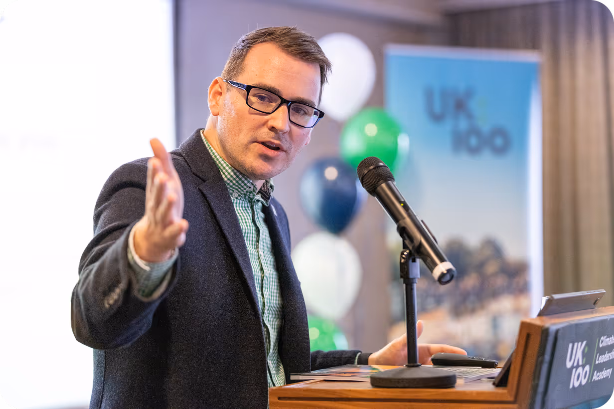 Speaker presenting at conference with microphone and UK 100 backdrop