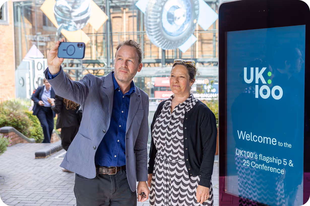 Two people taking a selfie at UK100 conference welcome sign
