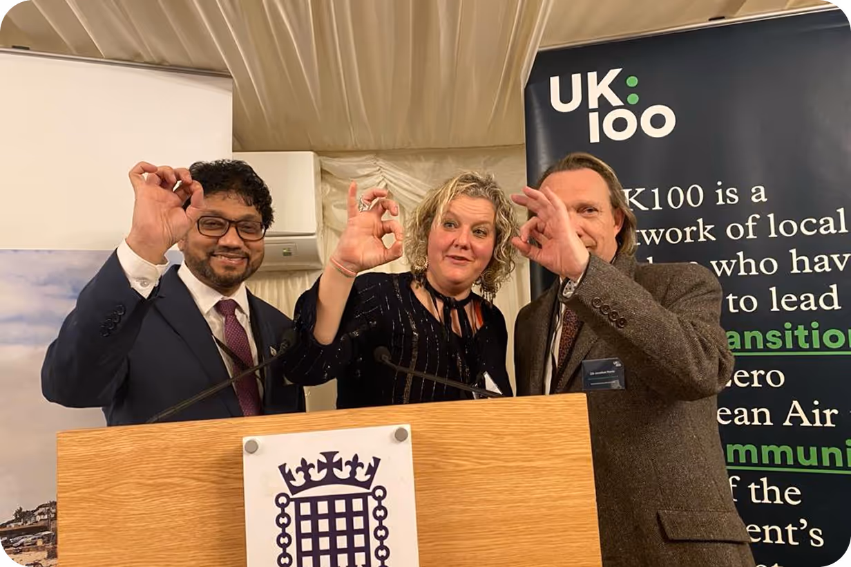 Three people posing with hand gestures at UK100 event podium