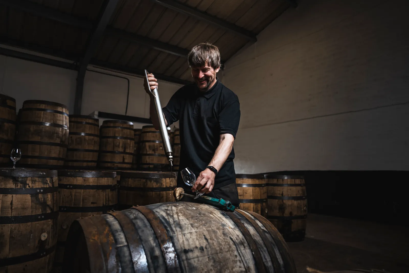 A man in a dark shirt extracting liquid from a wooden barrel with a metal tool into a glass inside a dimly lit warehouse filled with barrels.