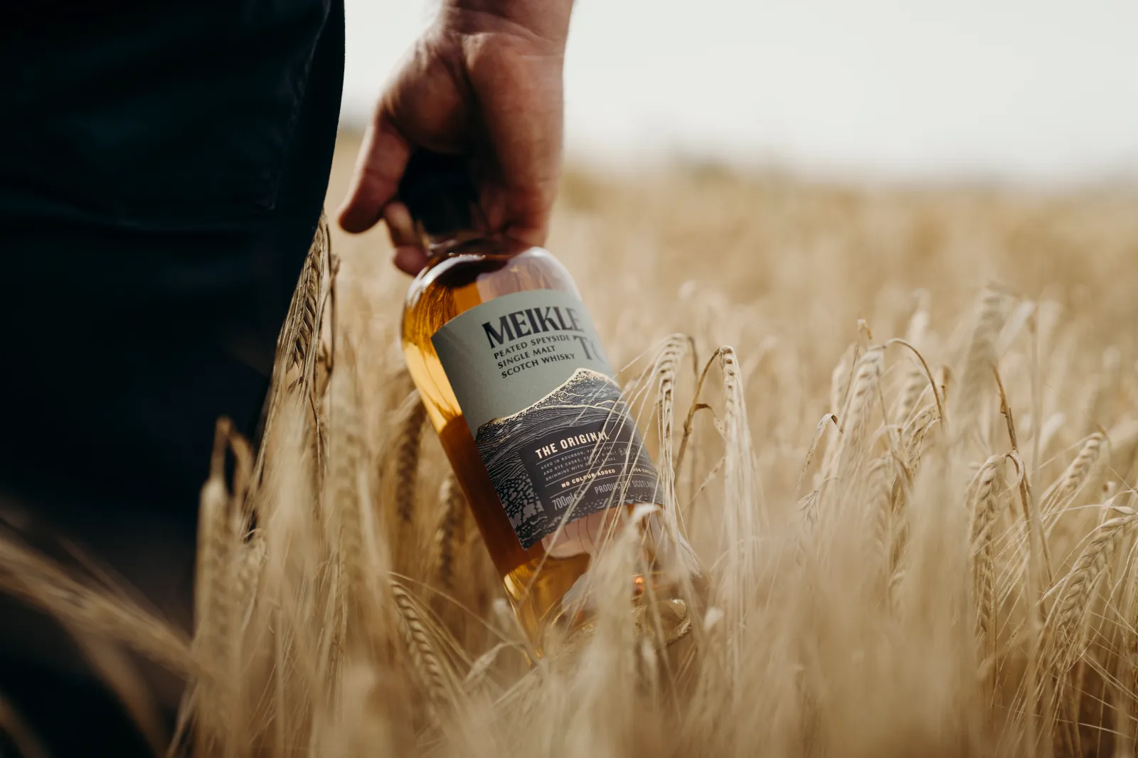 Hand holding a bottle of Meikle Toir peated Speyside single malt Scotch whisky in a golden wheat field.