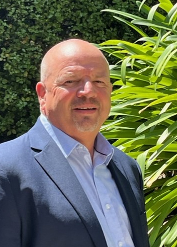 Middle-aged bald man wearing a navy blazer and light blue shirt standing outdoors near green leafy plants.