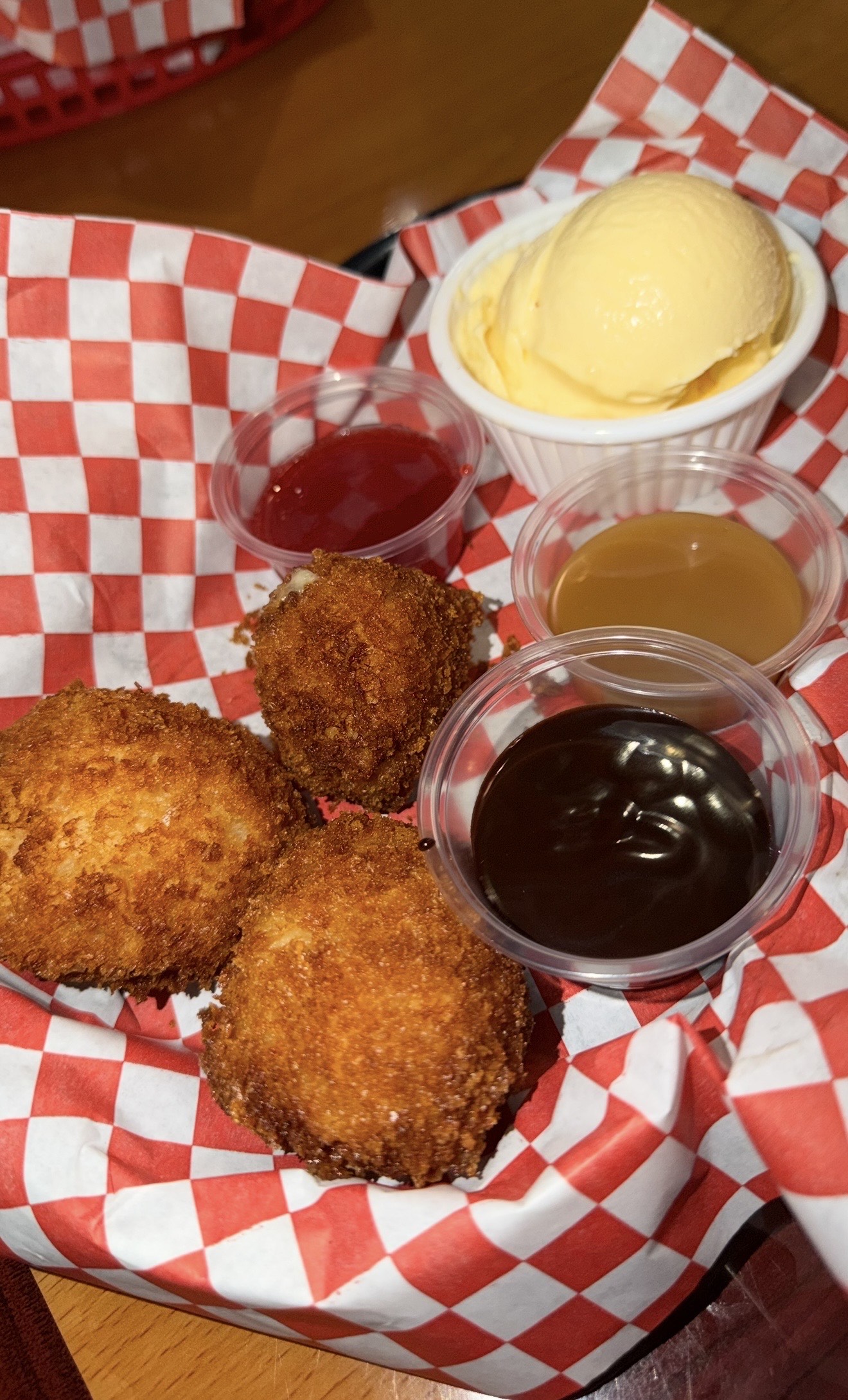 Three golden-brown, deep-fried treats sit on red checkered paper with vanilla ice cream and three sauce cups: berry, caramel, and chocolate.